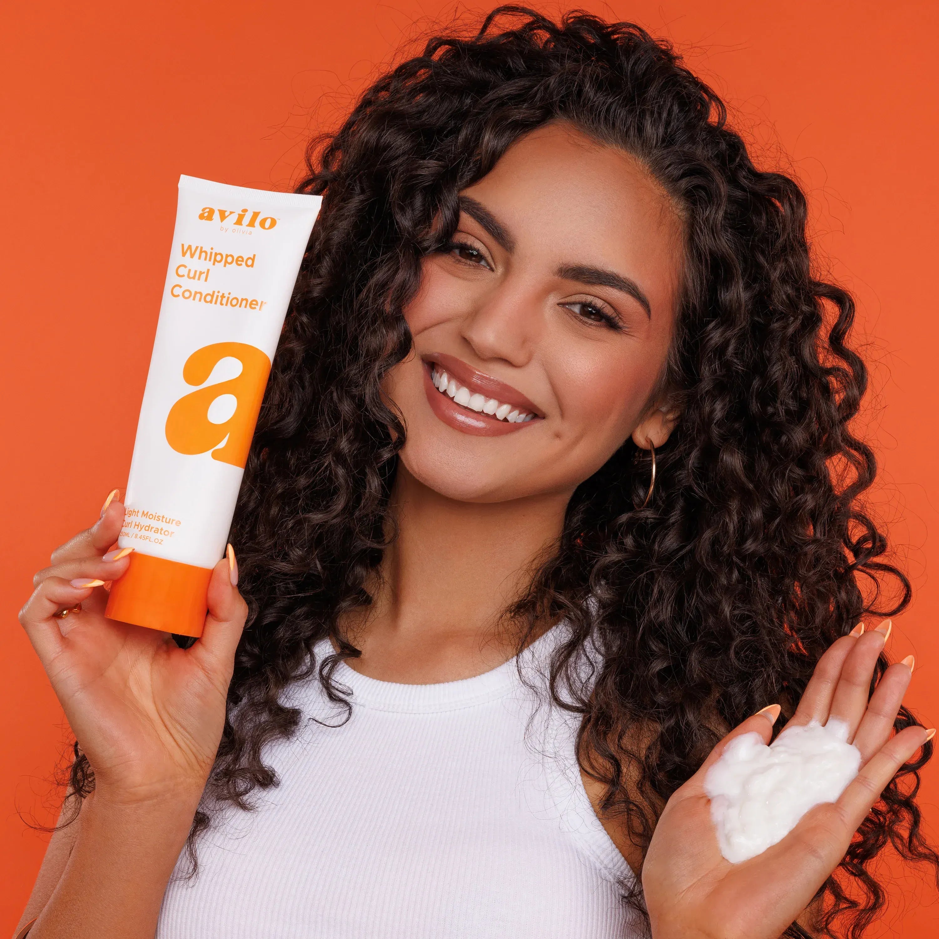 Woman with curly hair holding a bottle and a sample of Avliva Whipped Curl Conditioner against an orange background