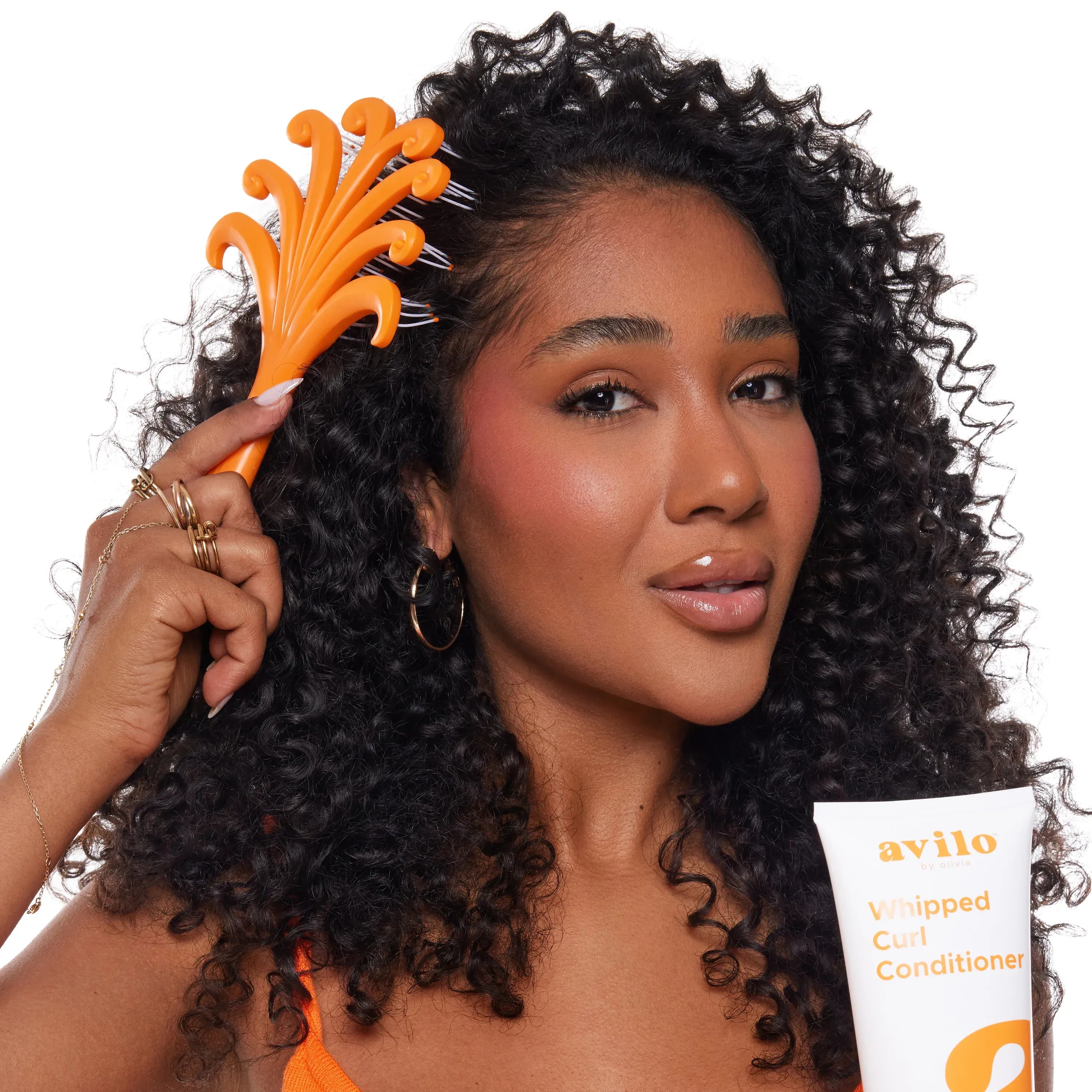 Woman with curly hair using an orange comb and holding a bottle of avilo whipped curl conditioner.