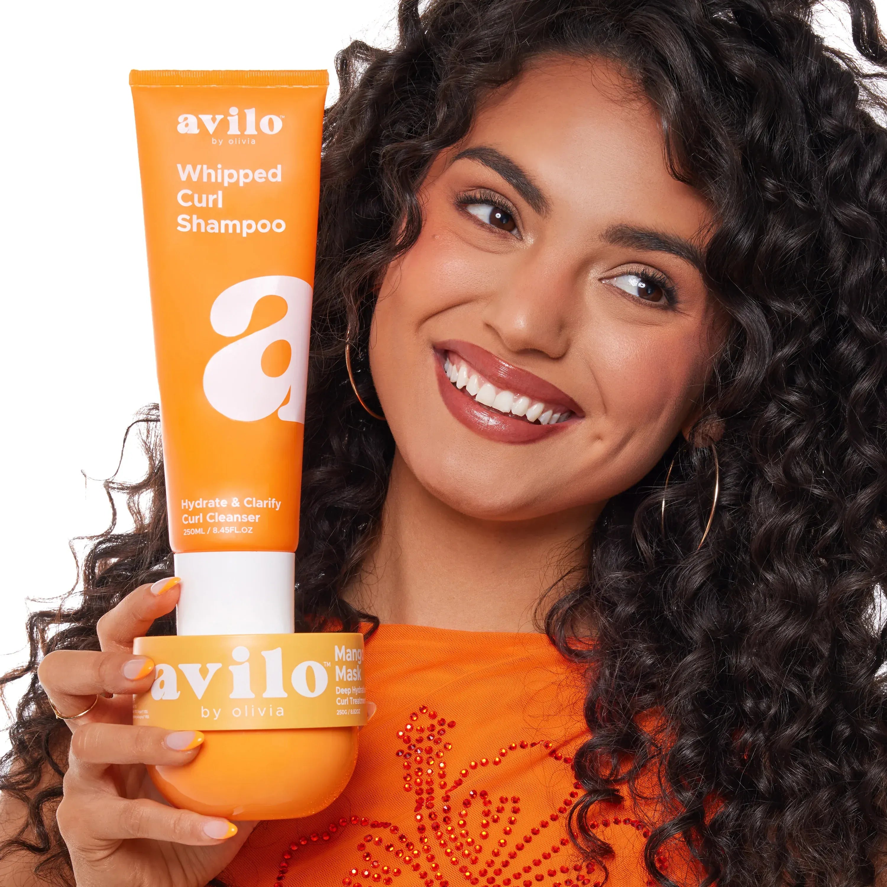 Woman with curly hair holding Avilo whipped curl shampoo and mask against a white background