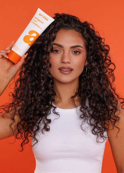 Woman with curly hair holding an Avlon product against an orange background

