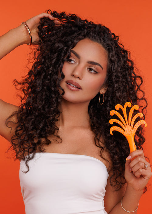 Woman with curly hair holding an orange hairbrush against an orange background