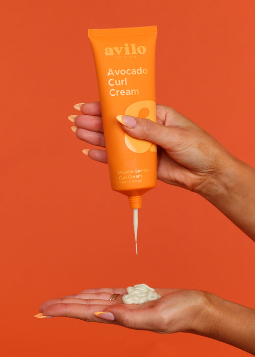 Hand holding an orange tube of Avilo Avocado Curl Cream against an orange background