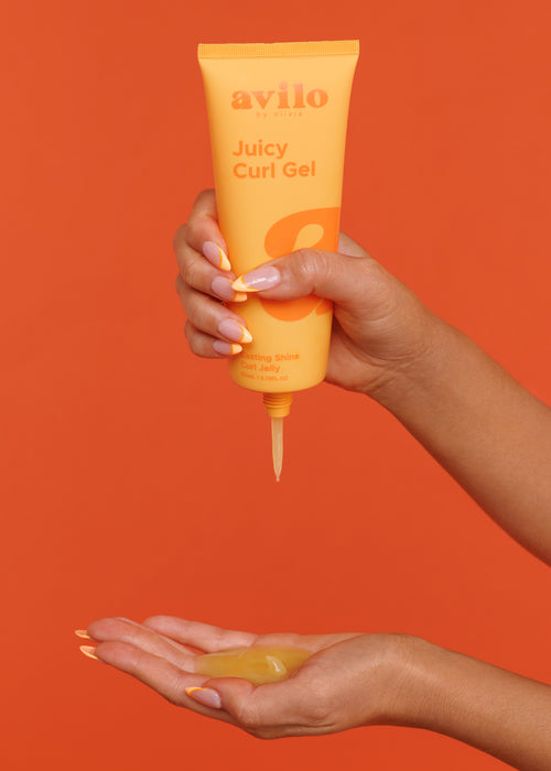 Hand holding a tube of avilo Juicy Curl Gel against an orange background

