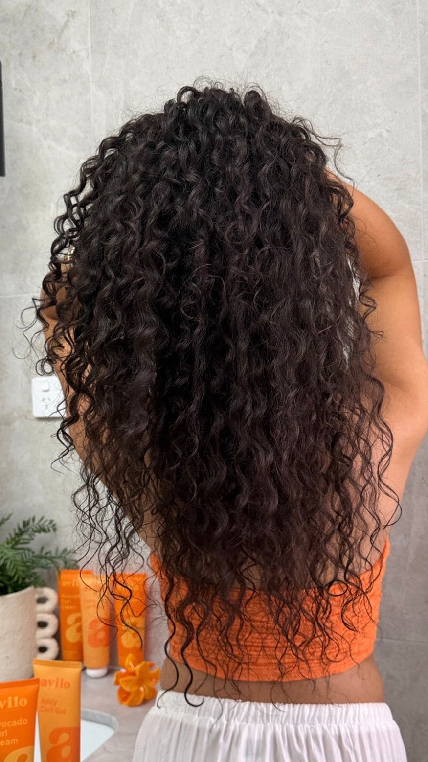 Person with long, curly hair in a bathroom setting with hair care products.
