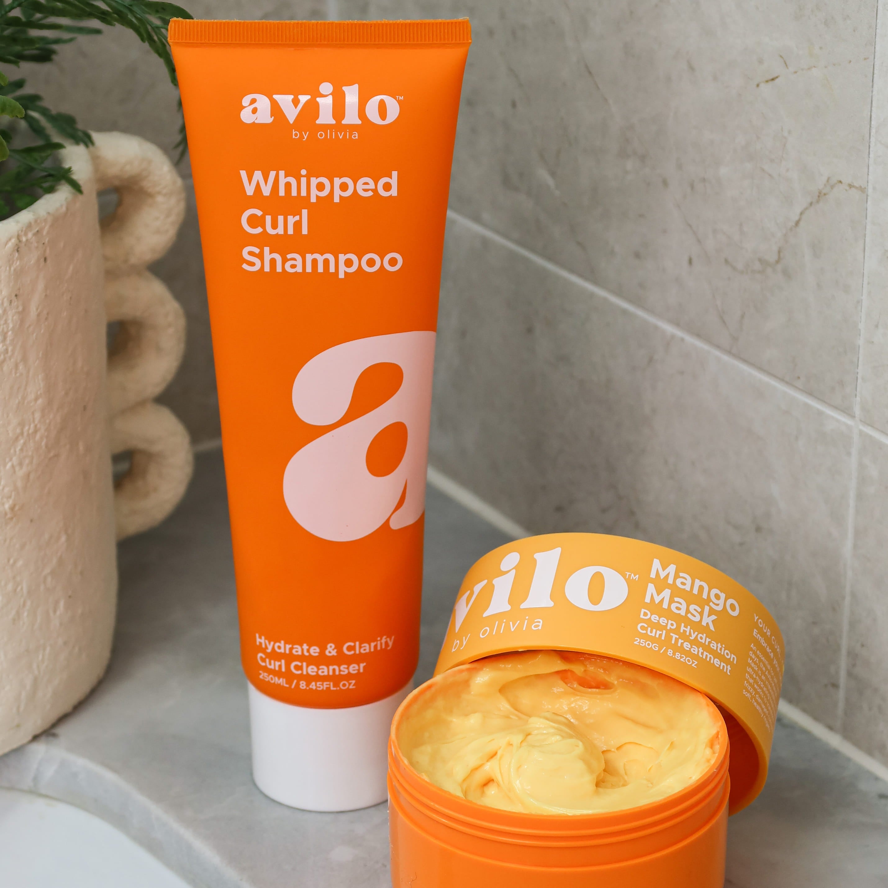 Avilo whipped curl shampoo tube and mango mask jar on a bathroom counter.