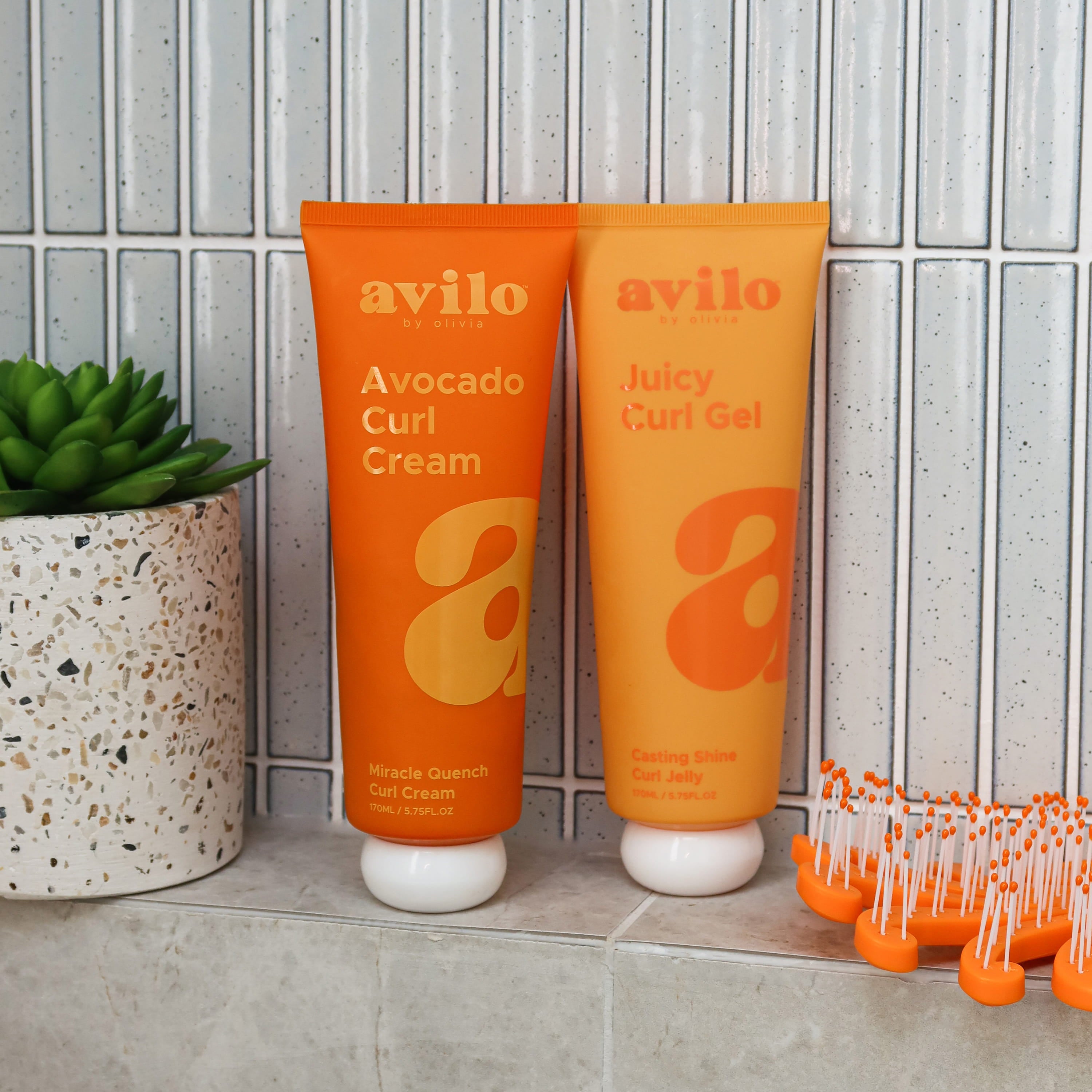 Two orange tubes of avilo hair care products on a tiled surface with a plant in the background.