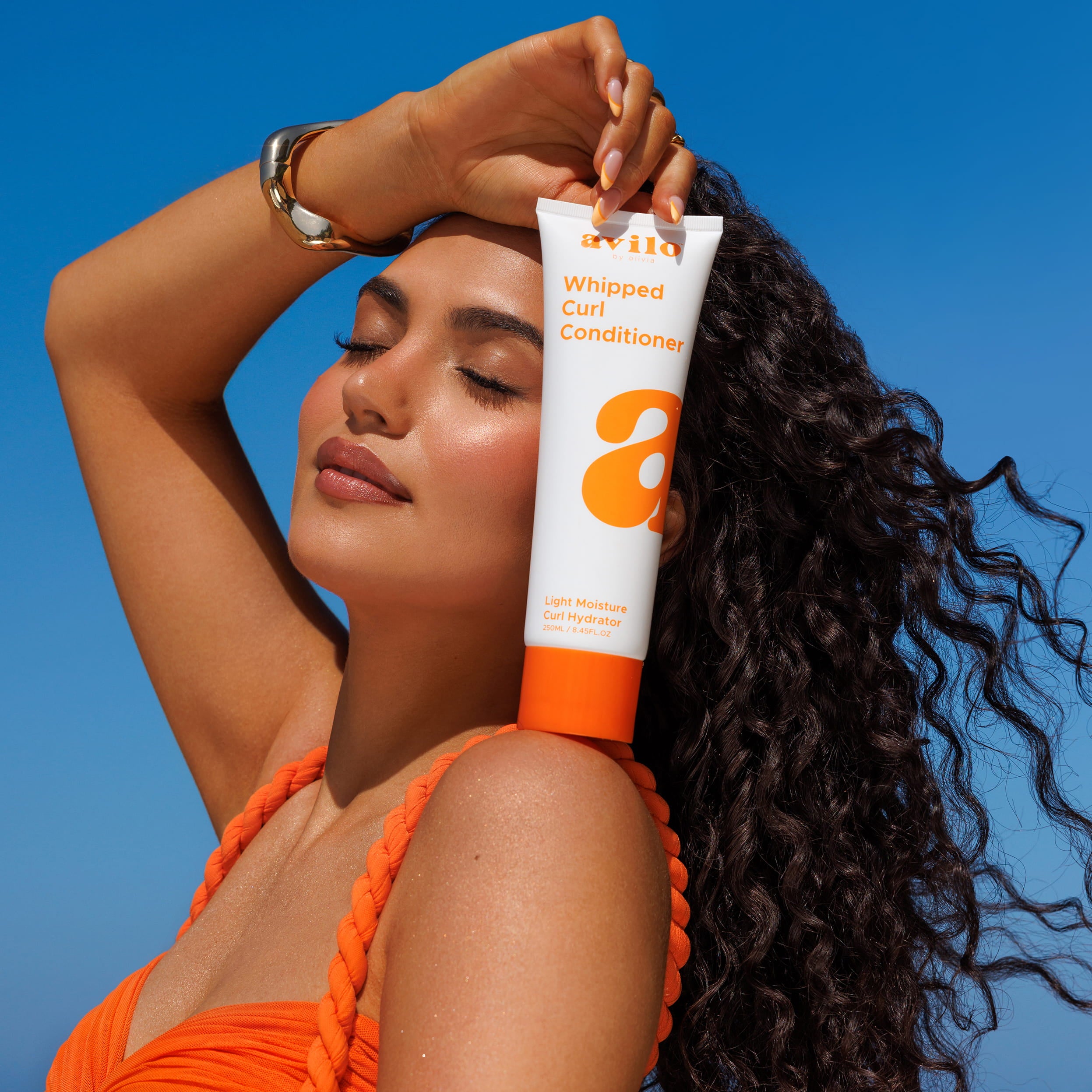 Woman holding a tube of 'a' Whipped Curl Conditioner against a blue sky