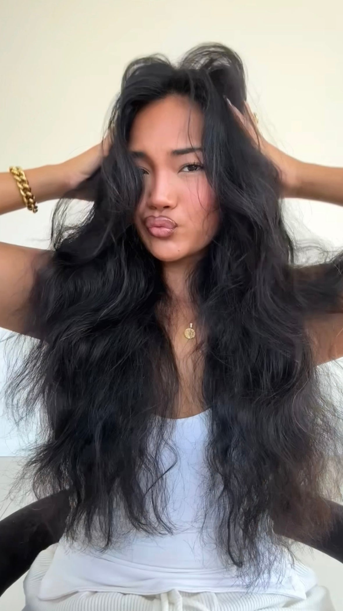 Woman with long, wavy black hair holding it with both hands against a neutral background
